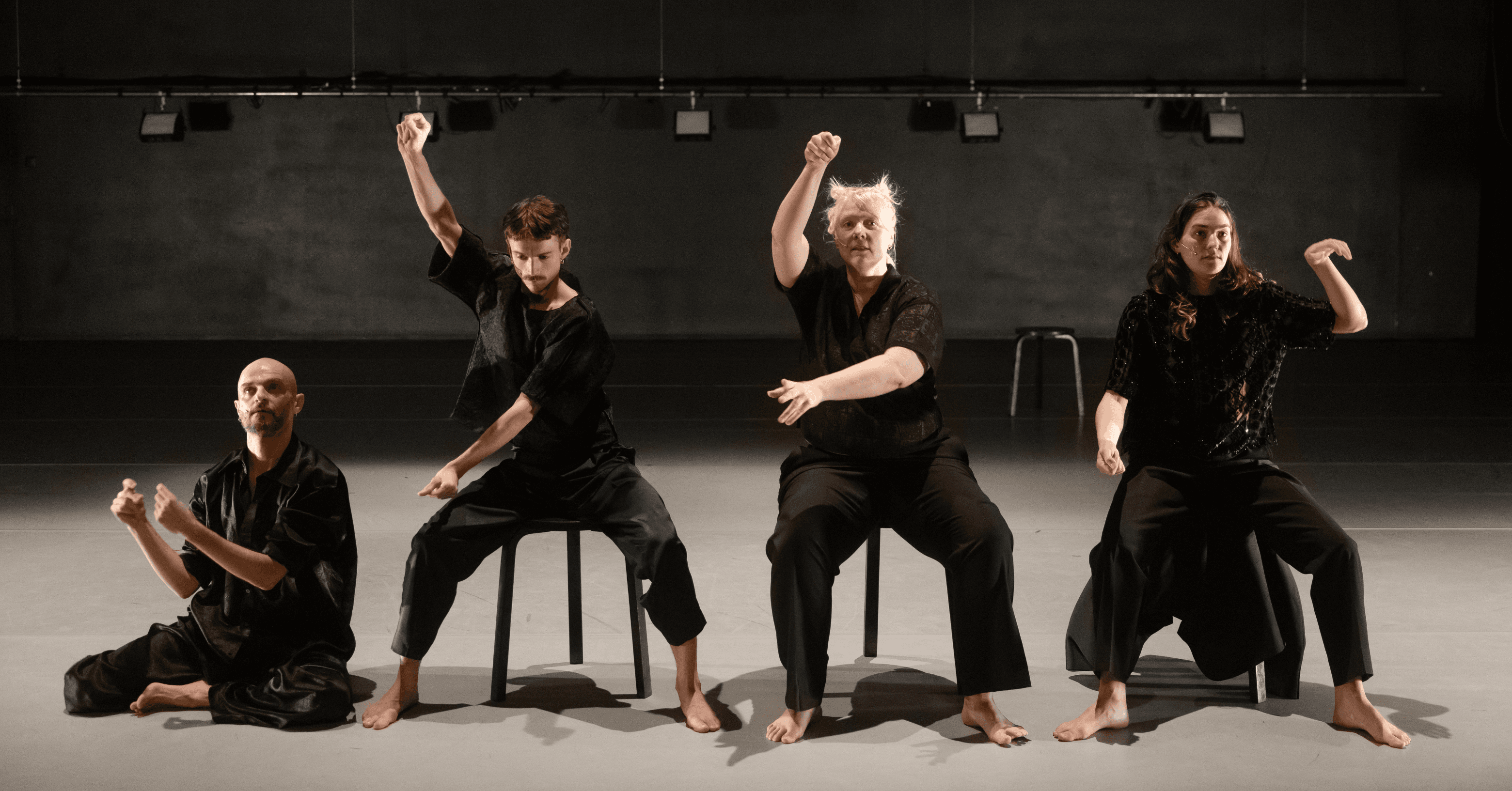 Four people dressed in black perform a seated dance routine on stage, each with unique arm gestures.