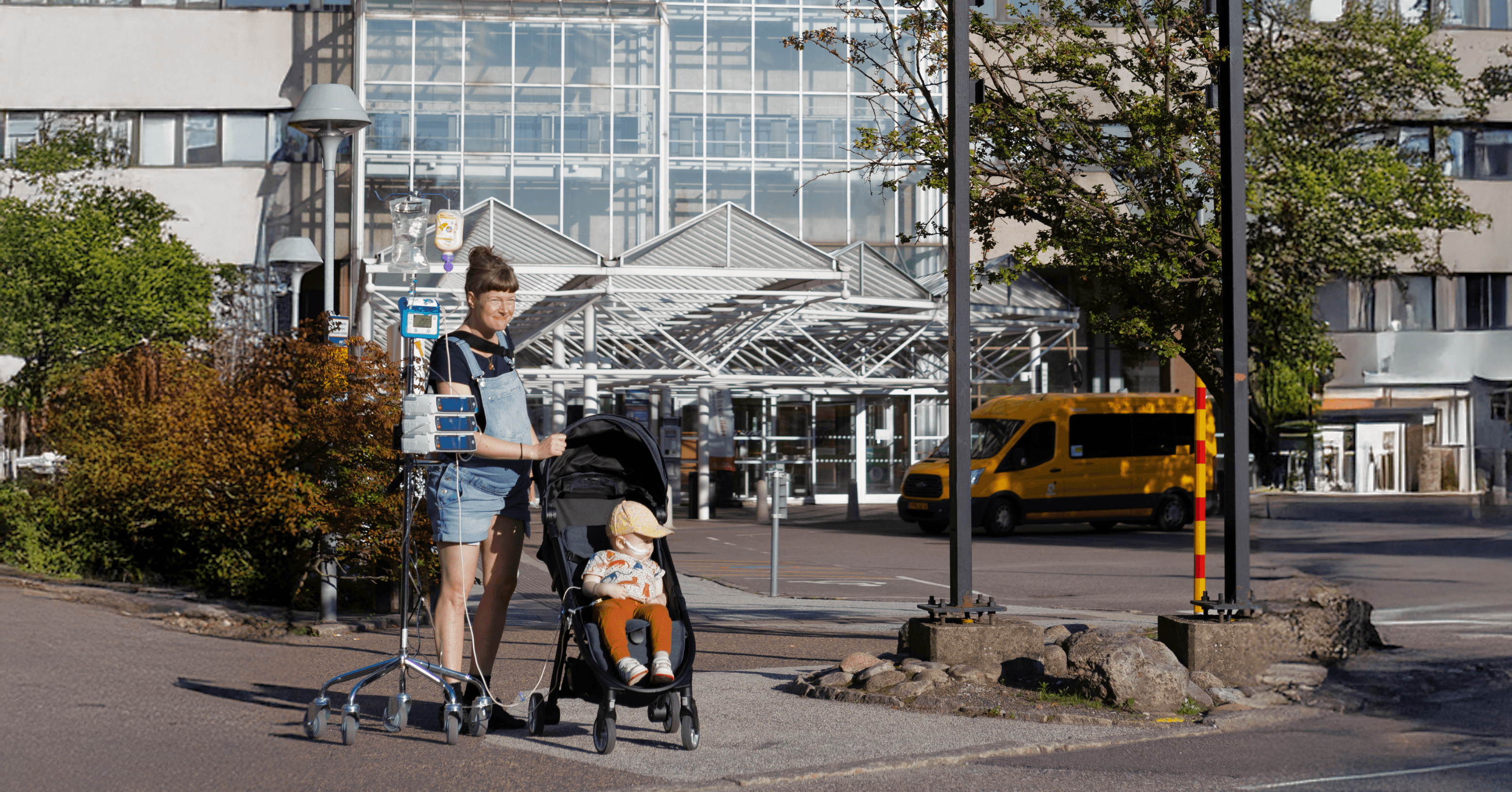 A person pushes a stroller with a child in it, accompanied by an IV stand, in front of a hospital entrance.