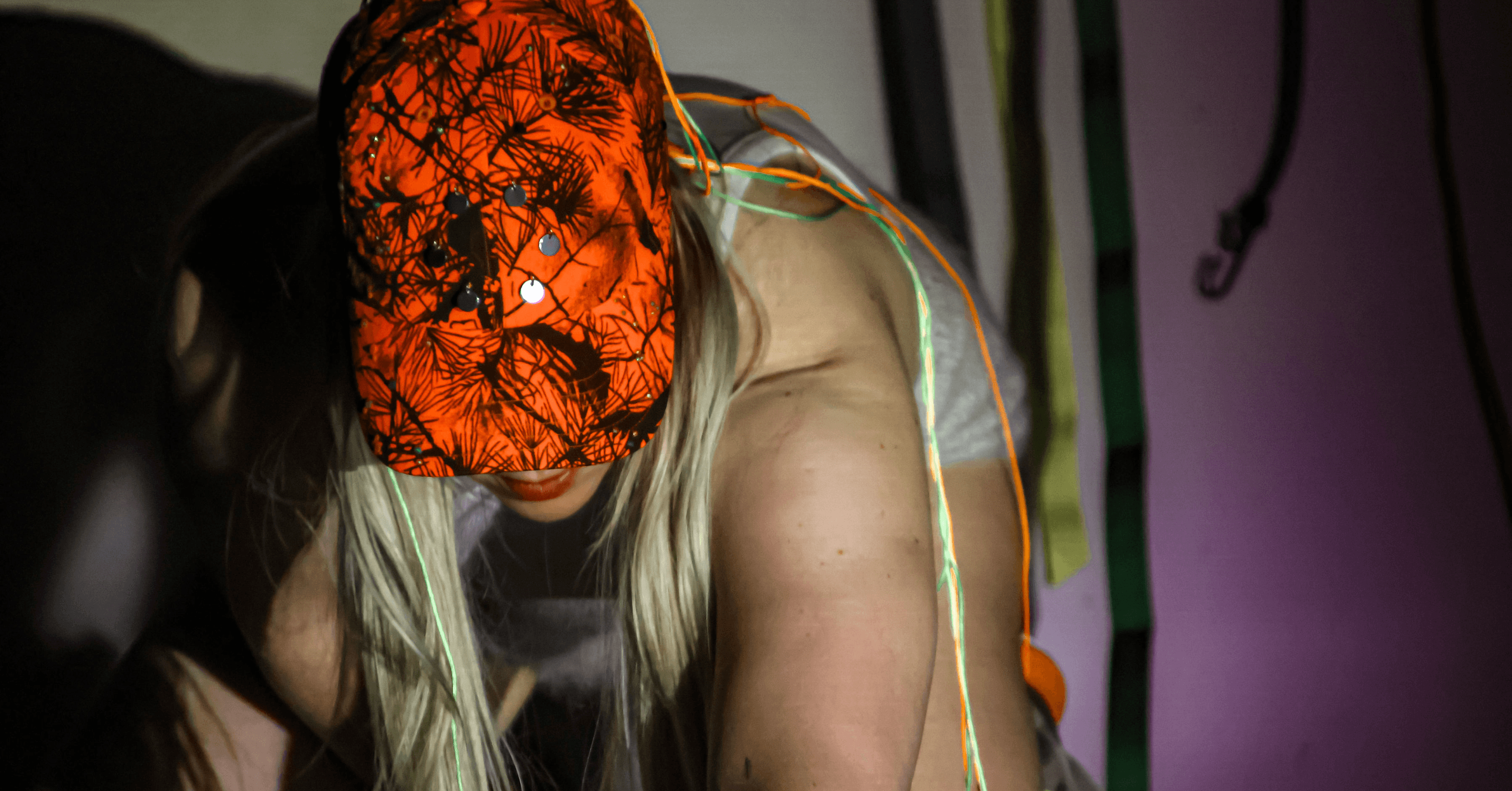 A person wearing a bright orange cap with a nature pattern and colorful wires draped over their shoulder is leaning forward.