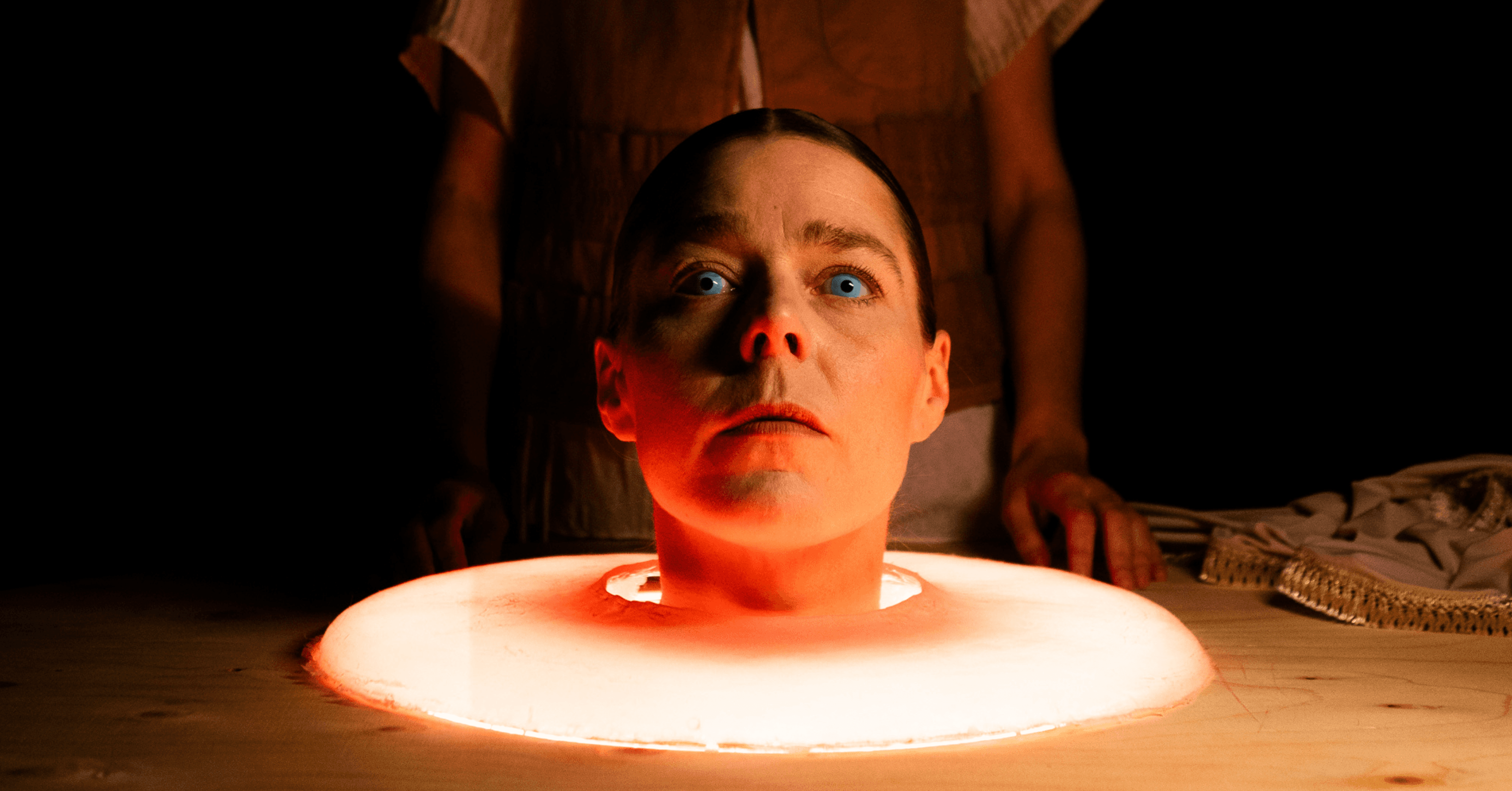A woman's head appears to be on a table surrounded by a glowing circular light, with someone standing behind her.