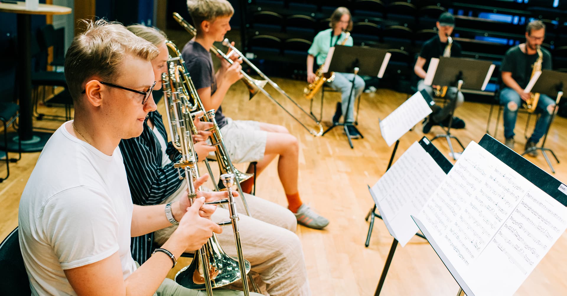 A group of musicians is playing brass and woodwind instruments, with sheet music on stands in front of them.