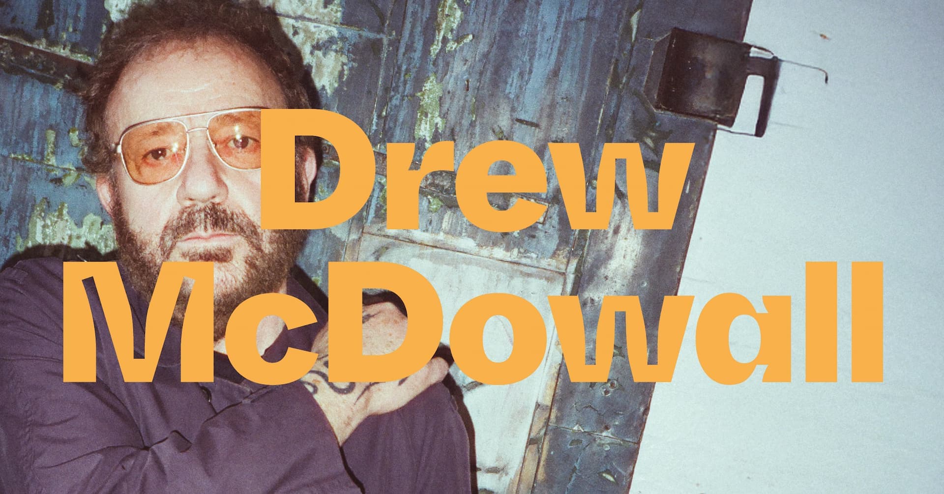 A man with a beard and glasses stands in front of a textured wall, with the name "Drew McDowall" in bold orange text across the image.