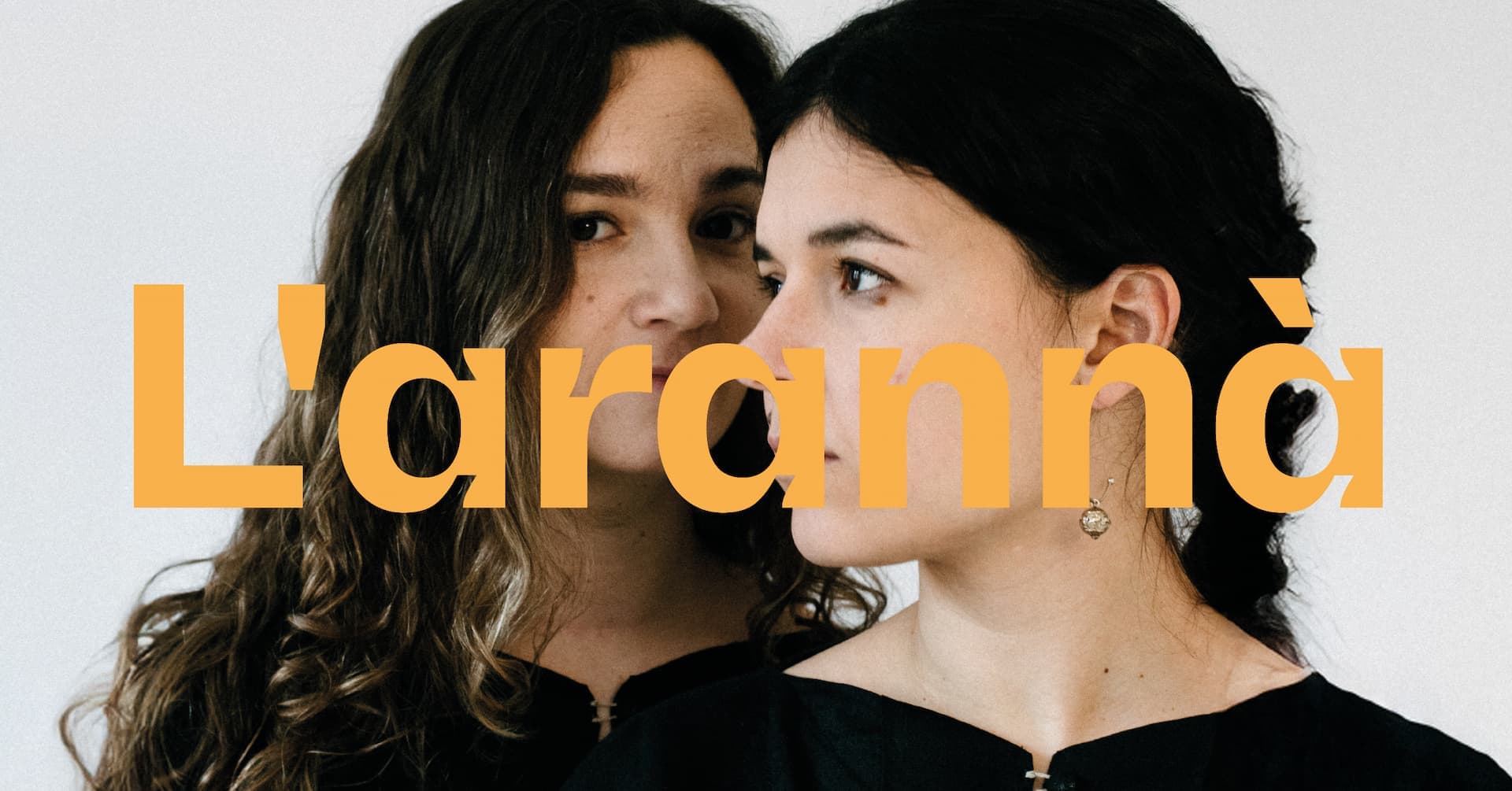 Two women in black attire are standing close together, with the text "L'arannà" overlaid in orange.